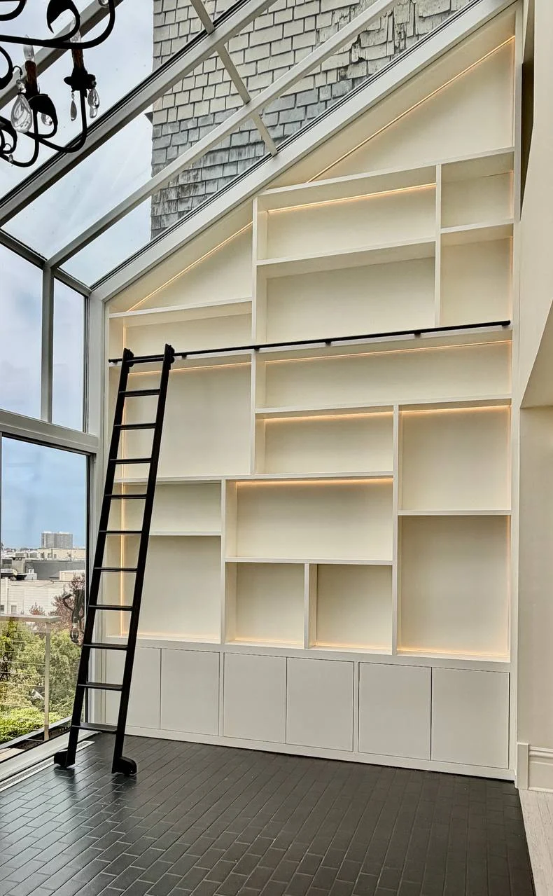 Tall lit library shelving beneath a sloping glass roof, with a dark rolling ladder